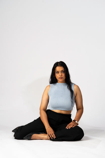 Person sitting cross-legged on a white background wearing a light blue sleeveless top and black pants.