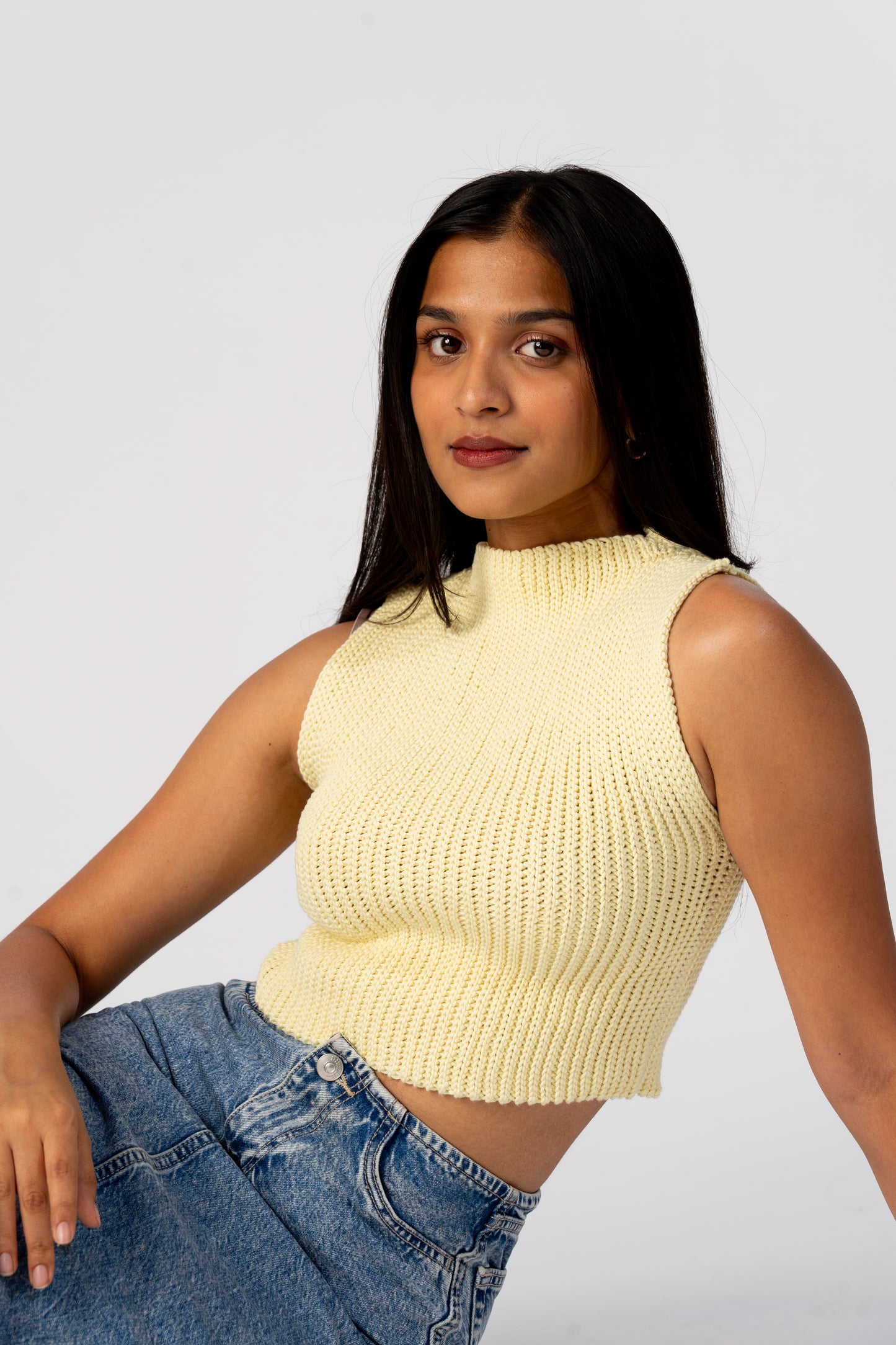 Woman wearing a light yellow crochet top and blue jeans against a white background