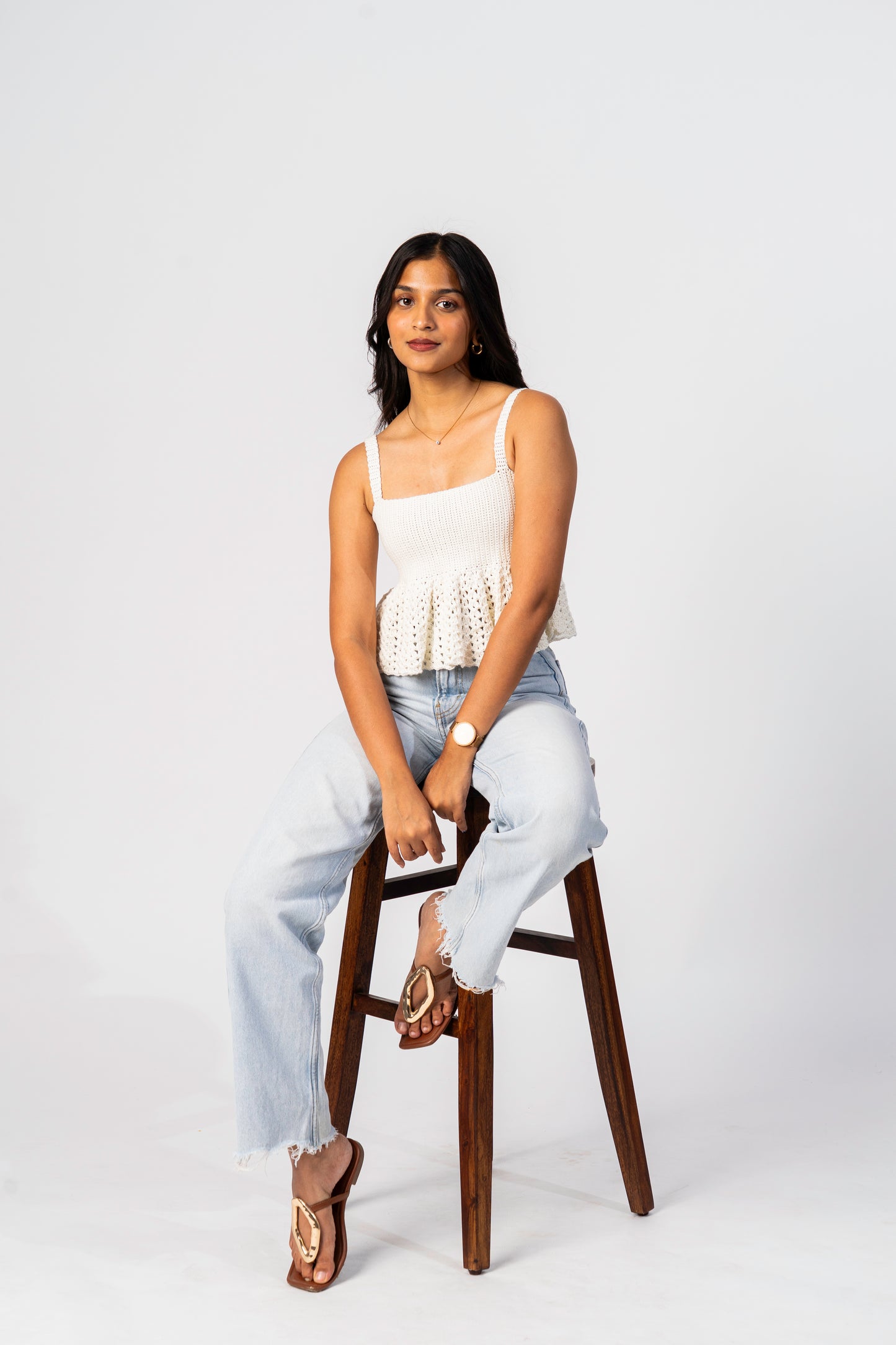 Woman sitting on a wooden stool wearing a white top and light blue jeans against a white background