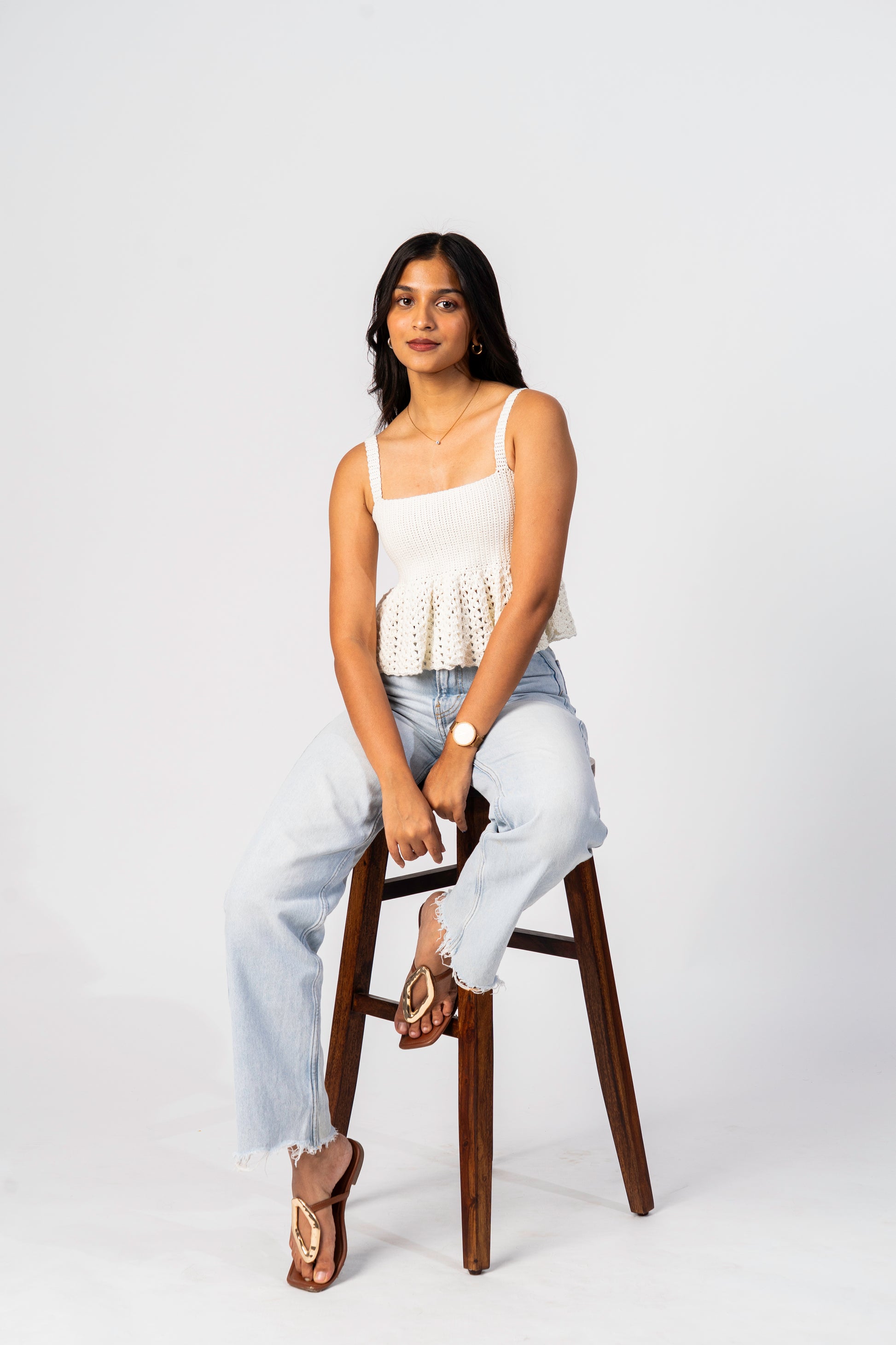 Woman sitting on a wooden stool wearing a white top and light blue jeans against a white background