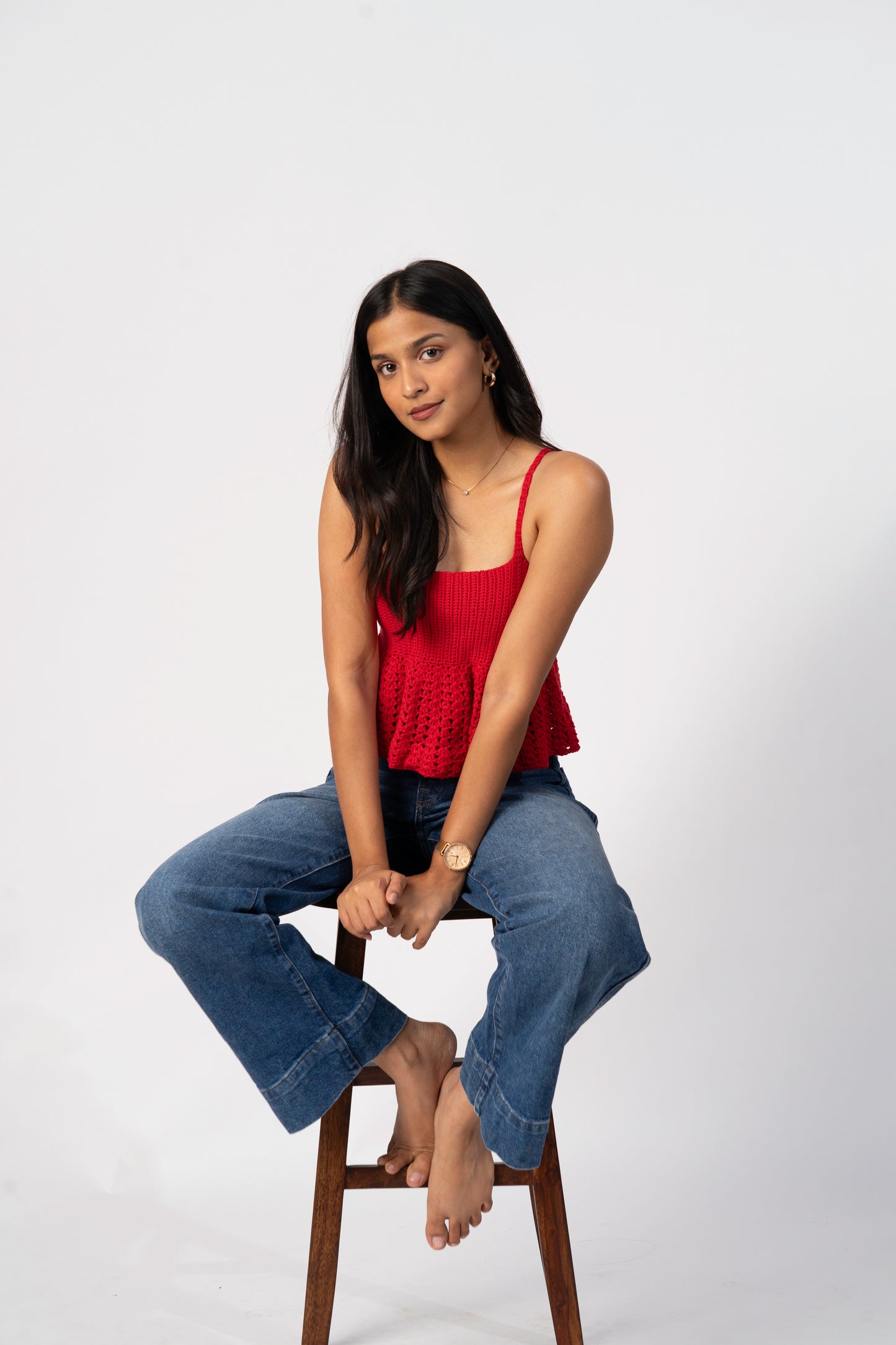 Woman sitting on a stool wearing a red top and blue jeans against a white background