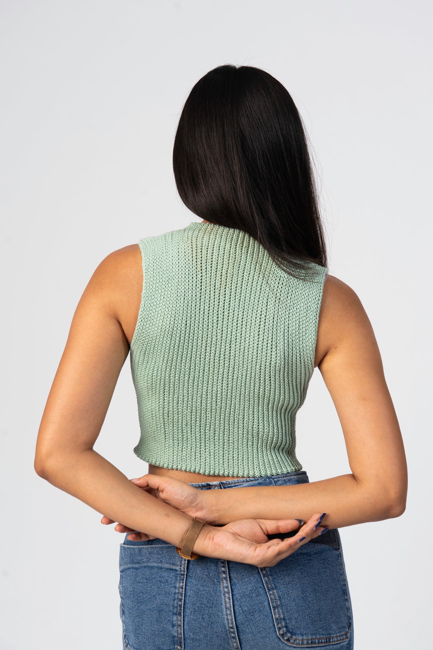 Person wearing a green crochet sleeveless top and blue jeans on a plain background