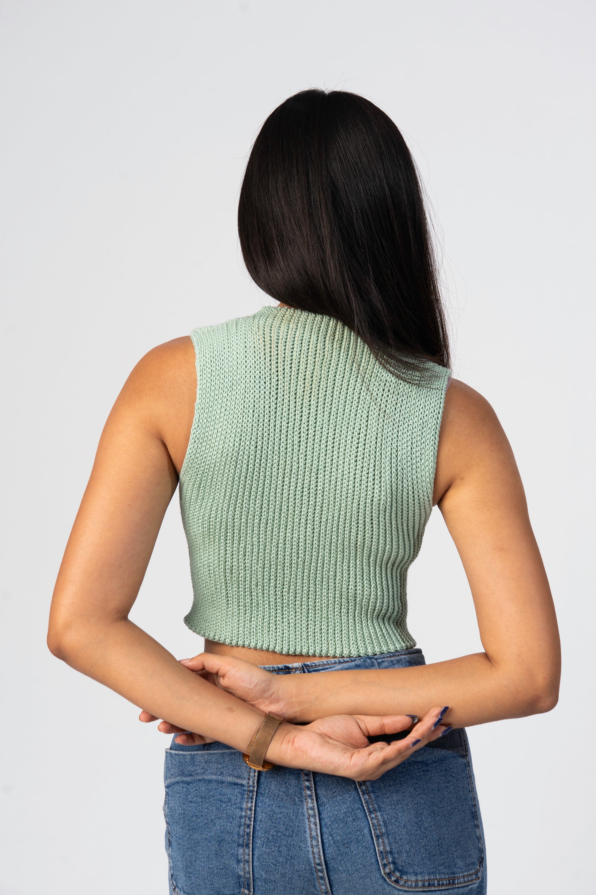 Person wearing a green crochet sleeveless top and blue jeans on a plain background